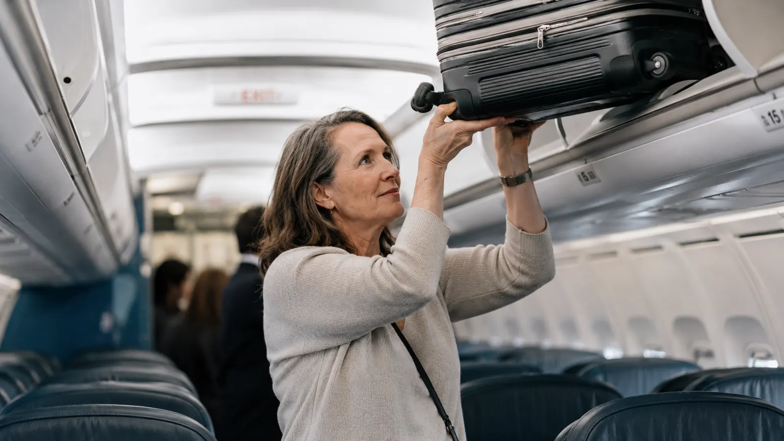 Woman in her 50s lifting a small carry-on roller bag into an airplane overhead bin with ease